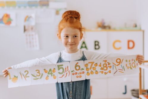 A young girl with red hair tied in a bun is standing in a classroom, smiling and holding a long strip of numbered cards. Each card displays a number from 1 to 10, illustrated with colorful fruits and vegetables. The background features a learning environment with alphabet letters on a shelf and various educational materials.